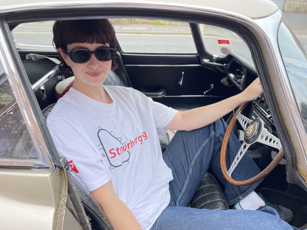 Holly at wheel of 1966 Jaguar E-type wearing a Stoürburgring t-shirt.